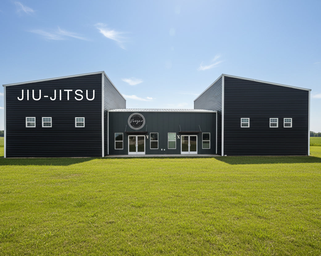 A large black metal building complex with two tall wings and a central shorter section, featuring white text reading 'JIU-JITSU' on the left wing and a circular logo on the center facade. The building is photographed outdoors against a pale sky with a light ground in front.